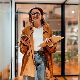 Smiling oung woman walking in front of office holding clipboard and smartphone